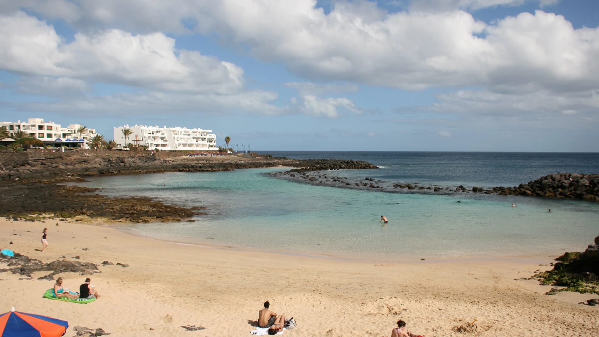 Playa del Jablillo in Costa Teguise, Lanzarote, a sheltered sandy cove with calm turquoise water behind a volcanic-rock breakwater and white apartment buildings