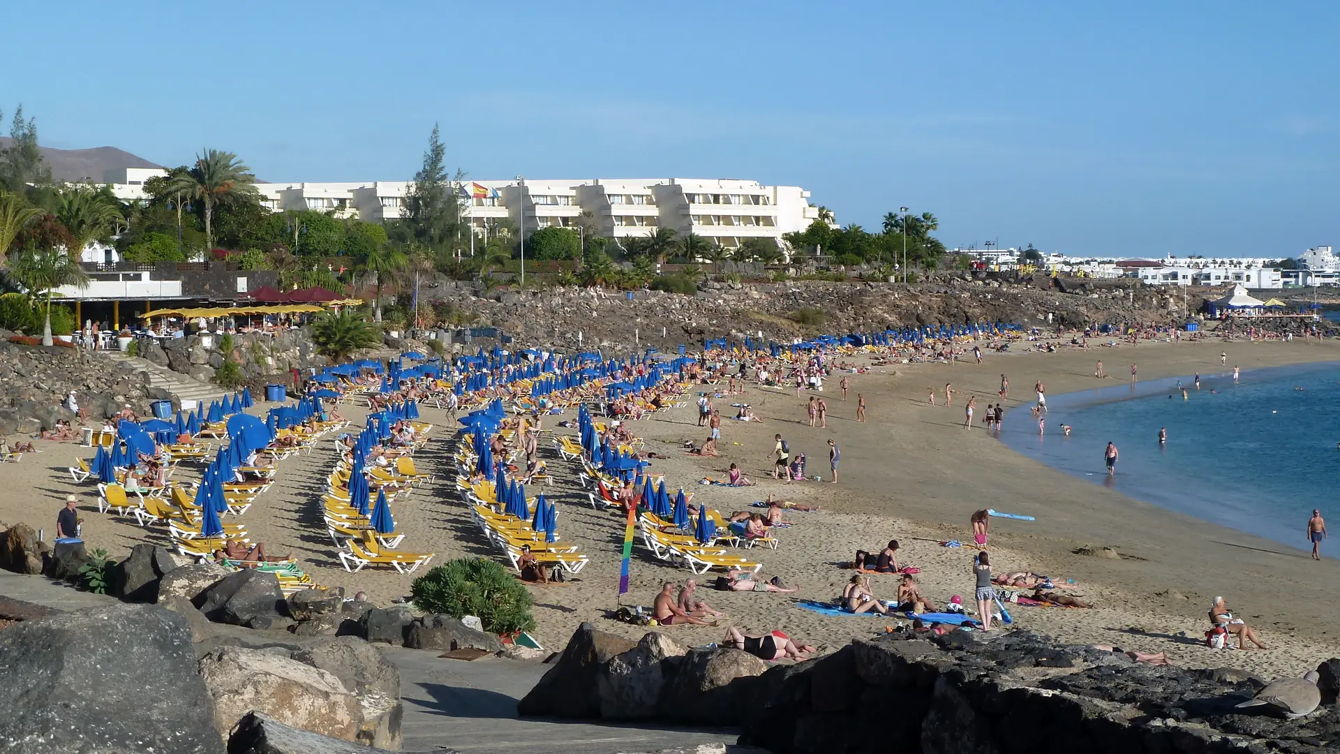 Playa Dorada in Playa Blanca, Lanzarote, a curved sandy beach lined with blue parasols and sunbeds below whitewashed resort buildings