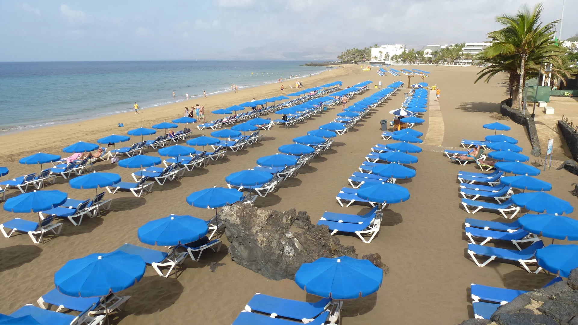 Rows of blue parasols and sunloungers on Playa Grande, Puerto del Carmen's main beach on Lanzarote, with palms and seafront hotels behind