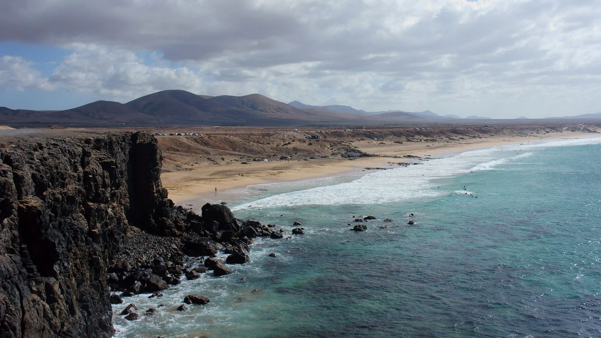 Winter sun on the beach at El Cotillo in north-west Fuerteventura, with surf rolling in below dark volcanic cliffs and brown hills behind