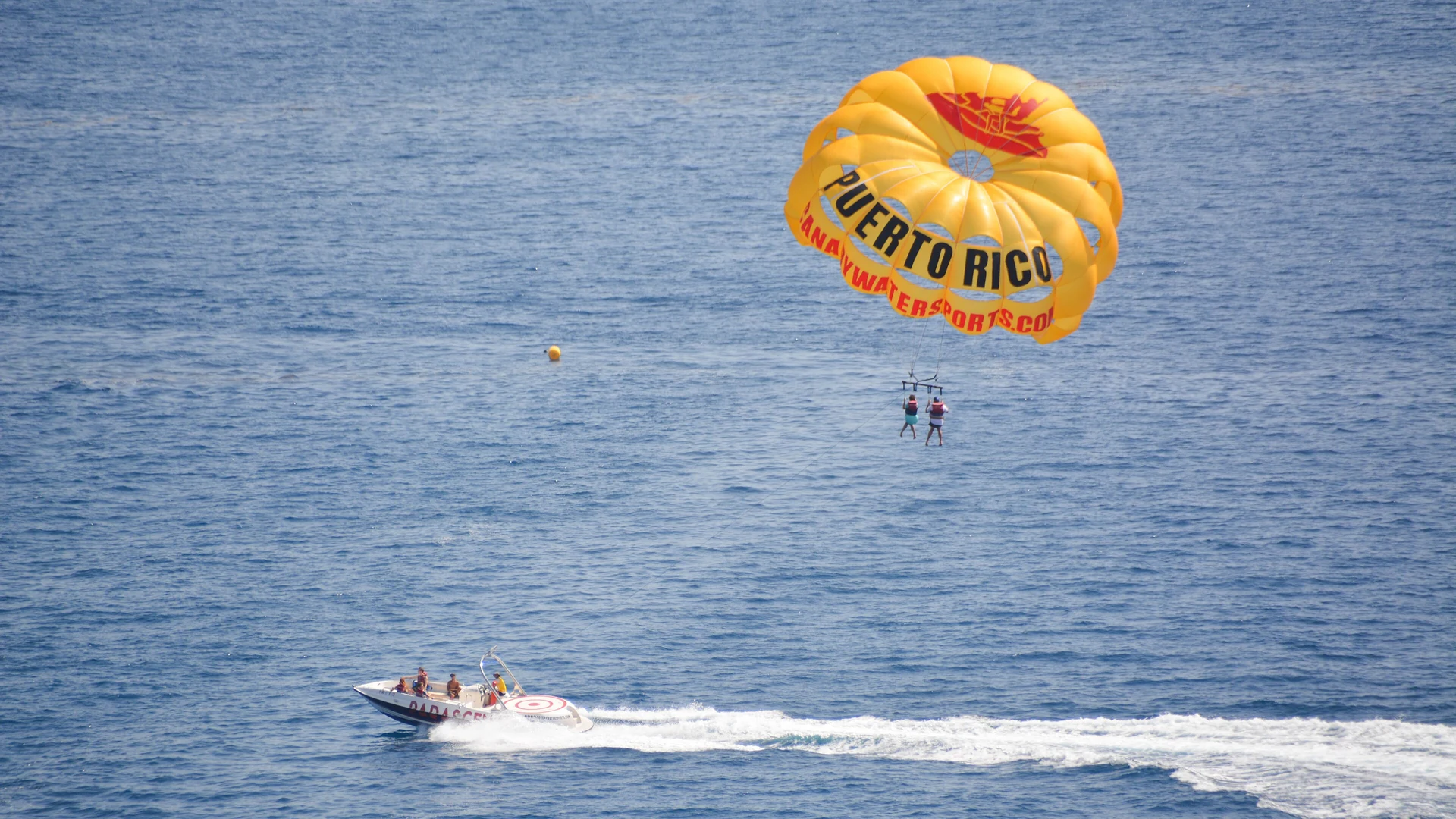 A yellow parasail with two people flying behind a speedboat over the sea off Puerto Rico, Gran Canaria