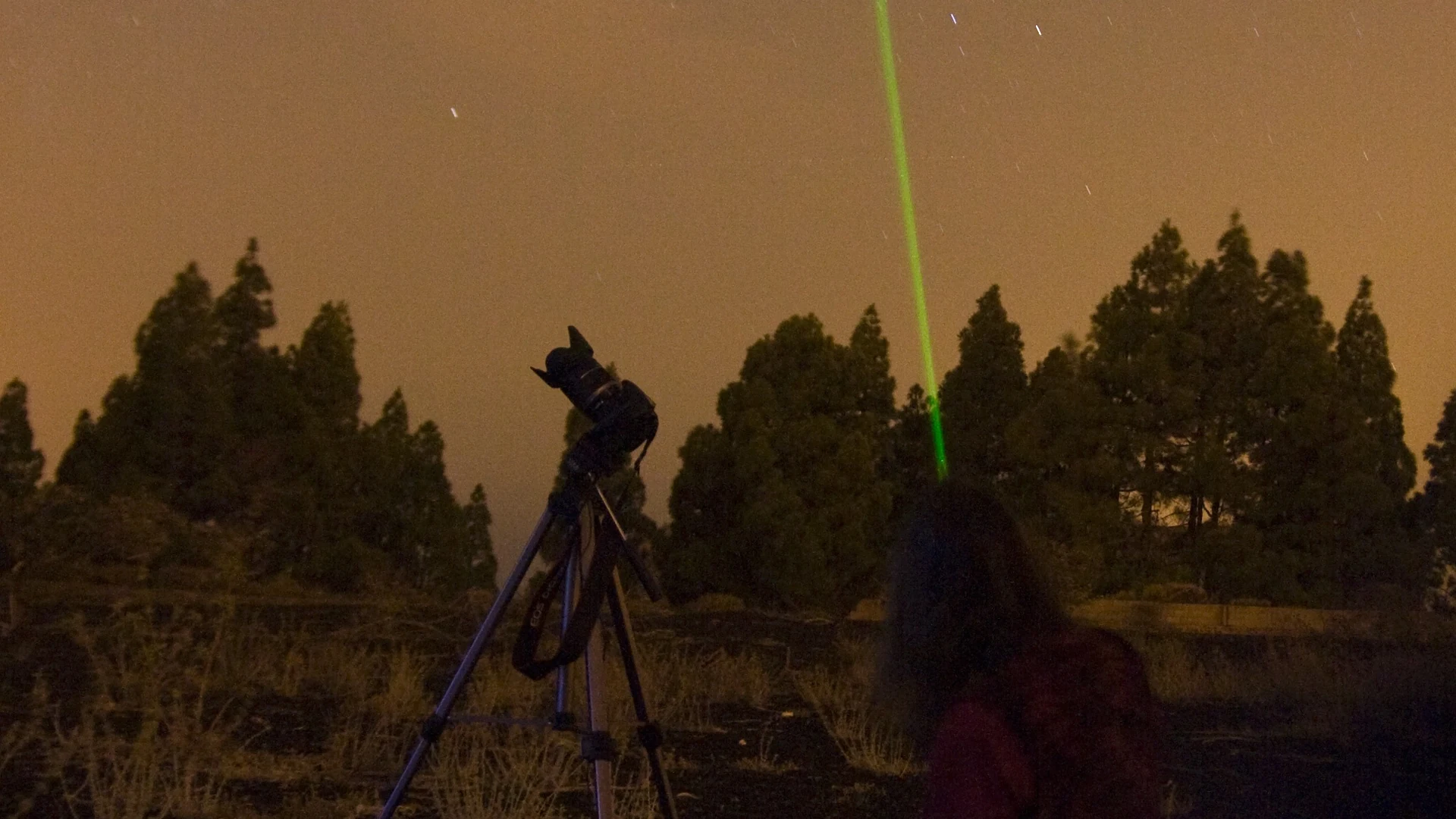 A green laser pointing at the stars above a pine forest, with a camera on a tripod, during a night-sky session in the mountains of Gran Canaria