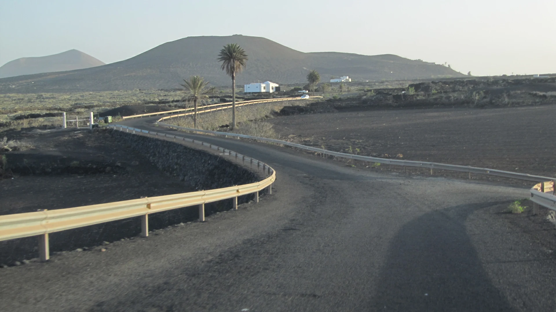 A paved road with crash barriers curving through black volcanic fields and palm trees towards a cinder cone on Lanzarote