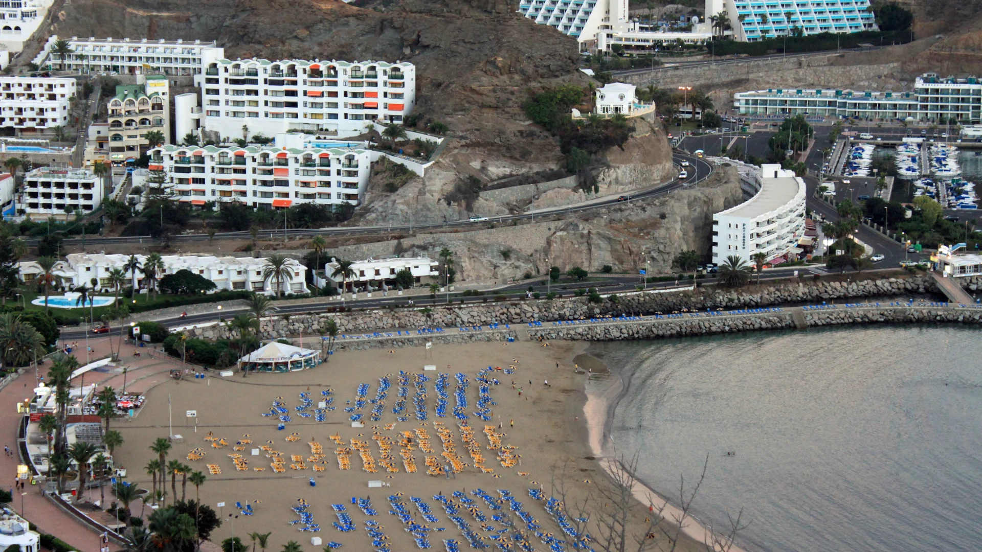 Puerto Rico beach in Gran Canaria from above, with rows of sunbeds, the breakwater and marina, and apartment blocks climbing the valley sides