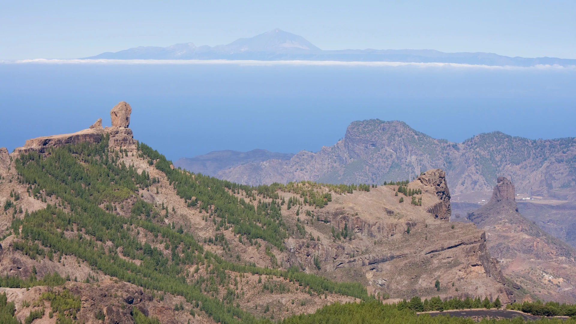Roque Nublo standing on its pine-covered ridge in central Gran Canaria, with Roque Bentayga to the right and Mount Teide rising above the clouds on the horizon
