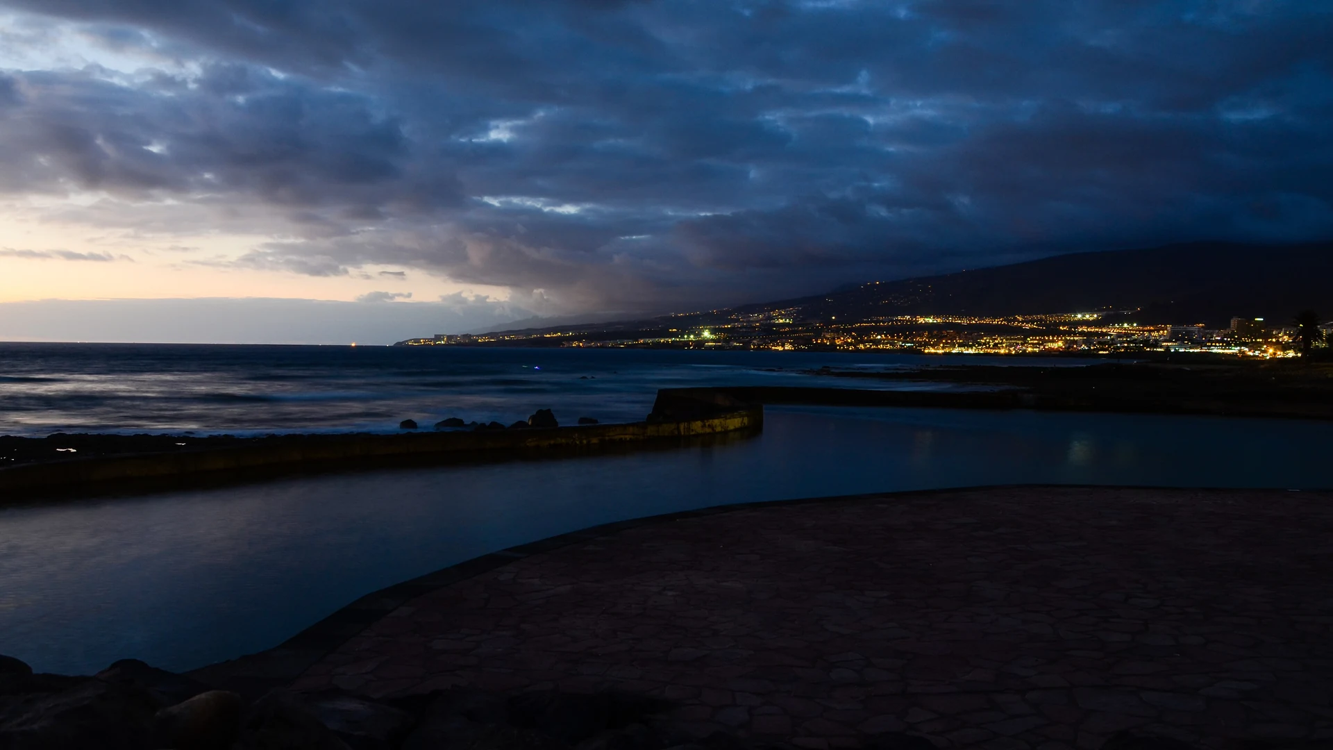 The lit resort strip along Tenerife's south-west coast at dusk, seen from the shoreline at Playa de las Américas